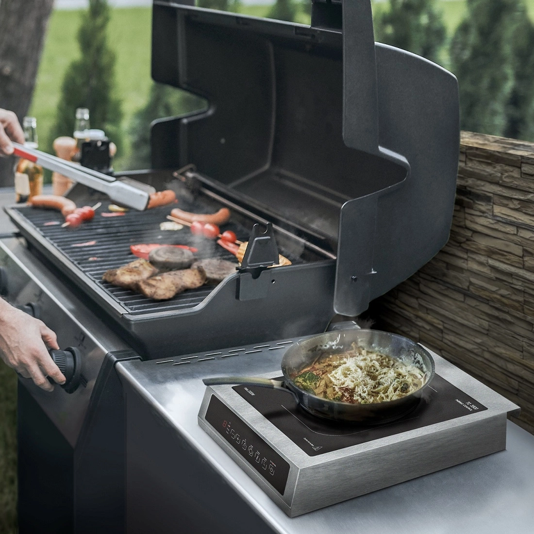 A man grills sausages and steaks on a gas grill, with an induction cooktop and pan next to it.