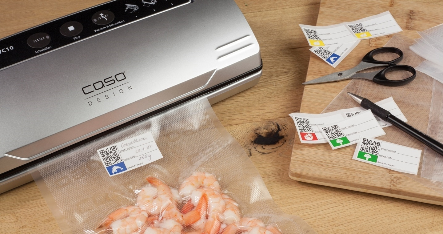Vakuumiergerät mit Garnelen, Etiketten und Zubehör auf Holzbrett.
