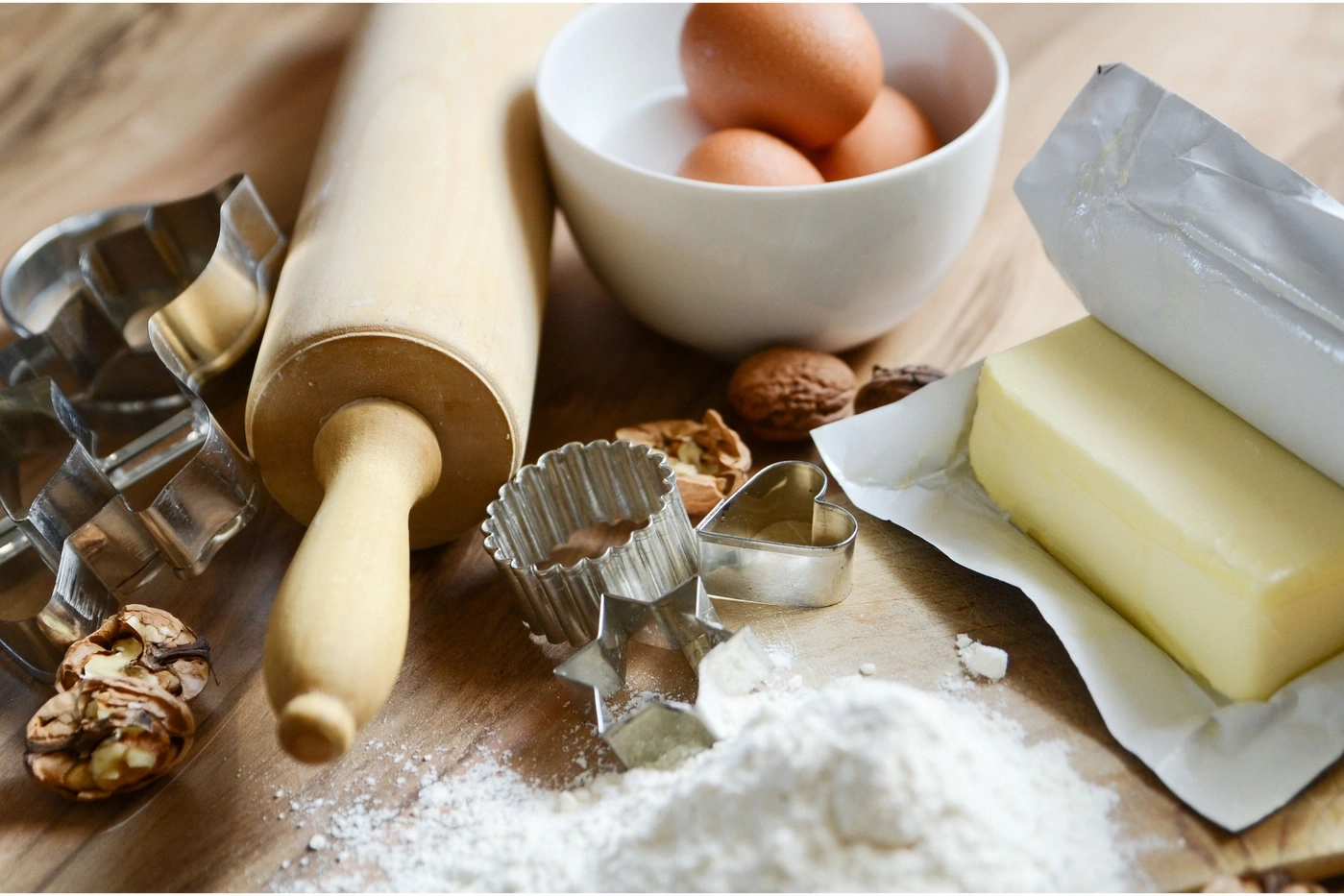 Baking ingredients on a wooden board: eggs, butter, flour, walnuts, cookie cutters, and rolling pin.