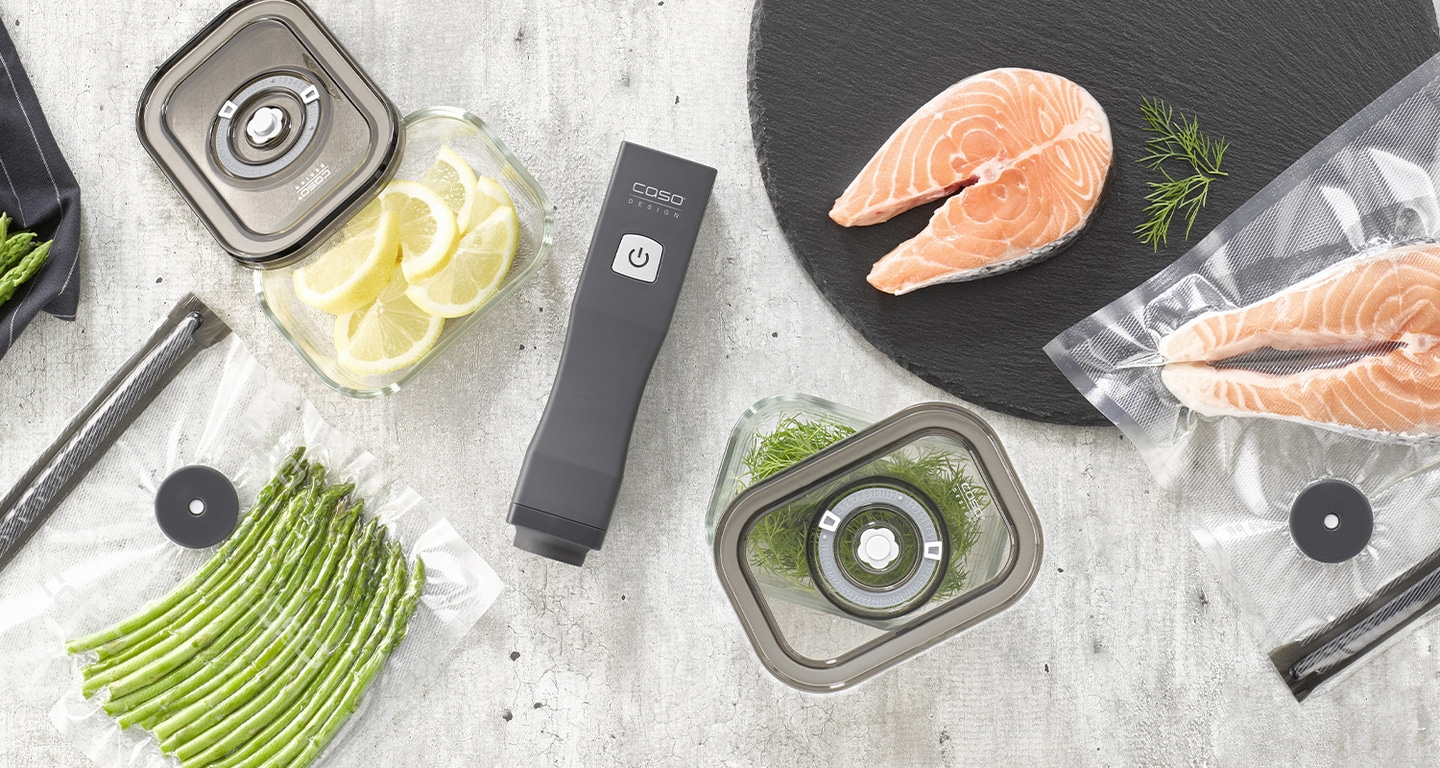 Vakuumiergerät mit Lebensmitteln wie Lachs, Spargel und Zitronen in Gläsern und Beuteln.