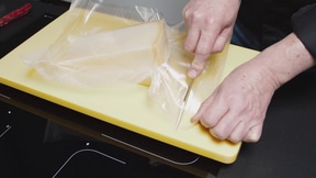 Person schneidet Käse aus der Verpackung auf einem gelben Brett mit einem Messer.