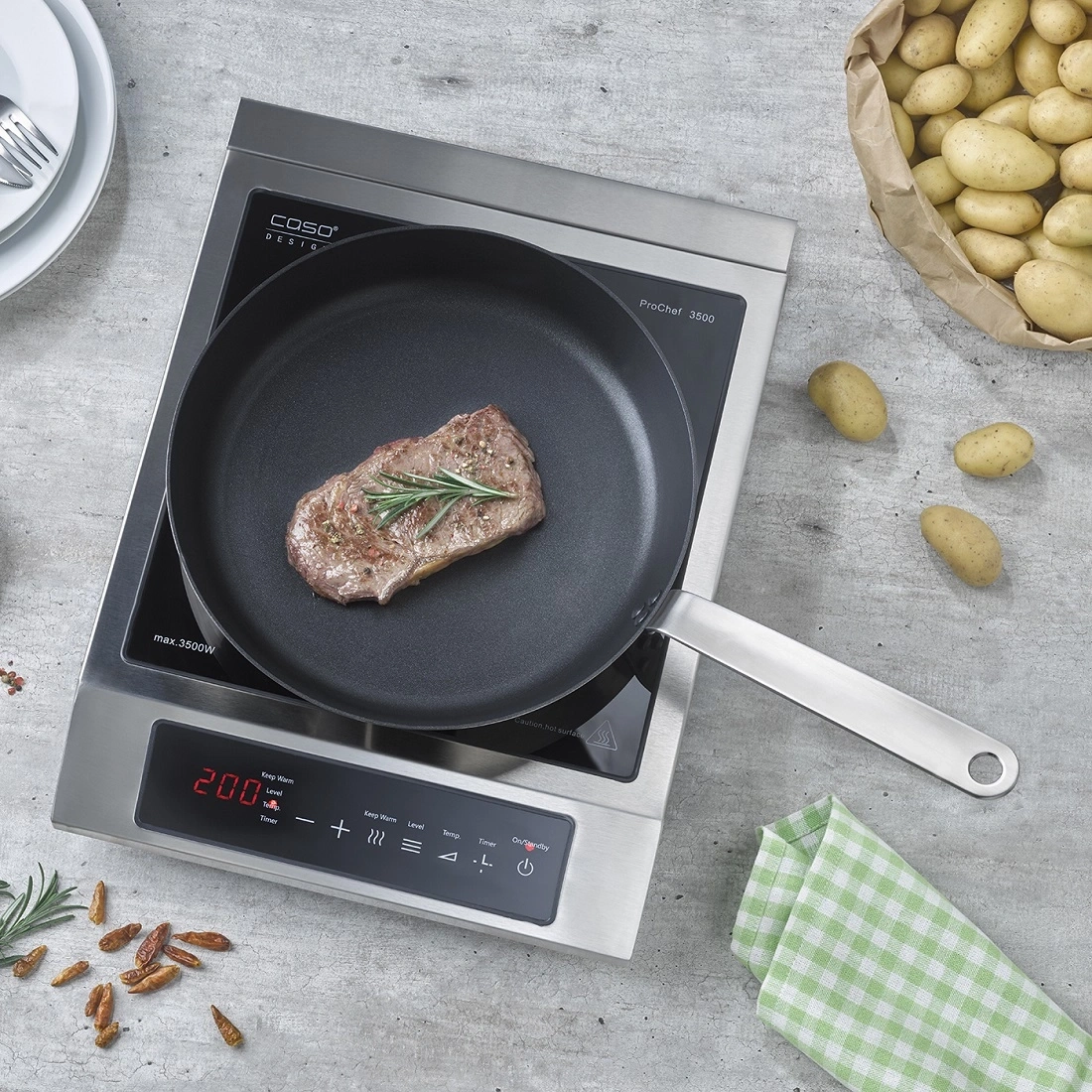 A steak fries in a pan on an induction hob, with potatoes nearby.