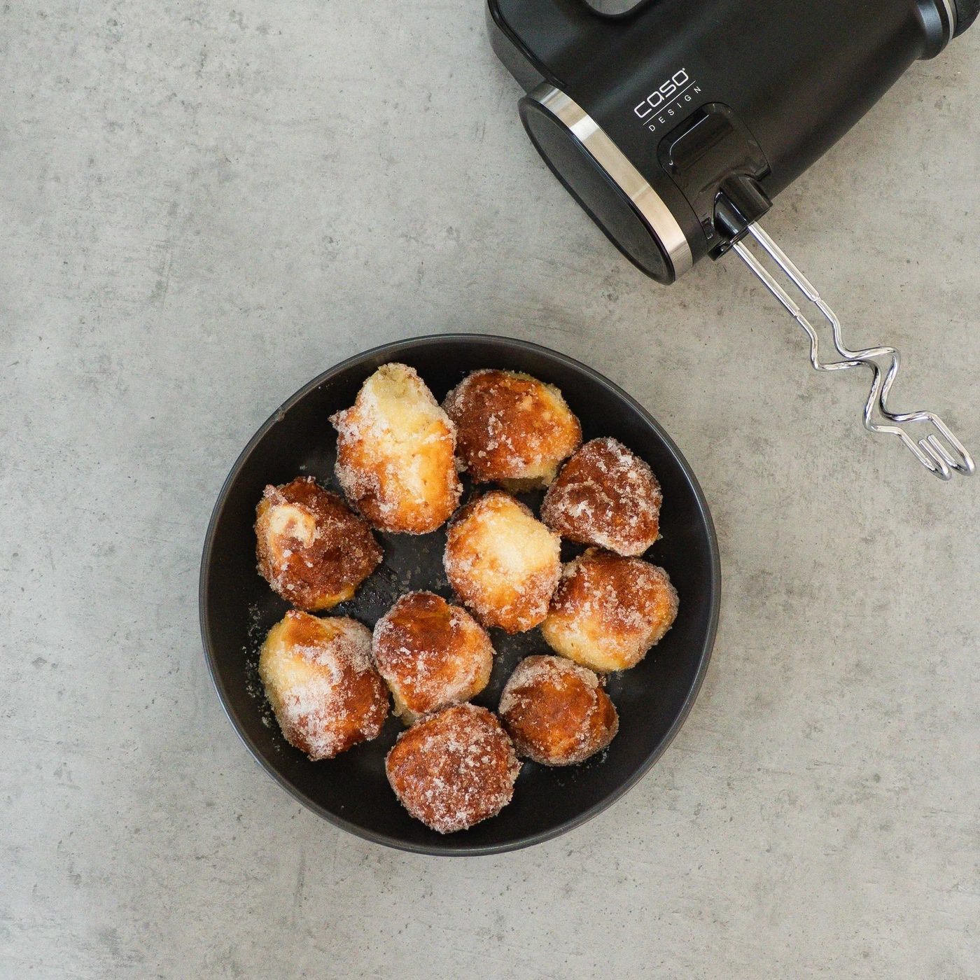 Ein Teller mit süßen, gezuckerten Krapfen neben einem Handmixer.