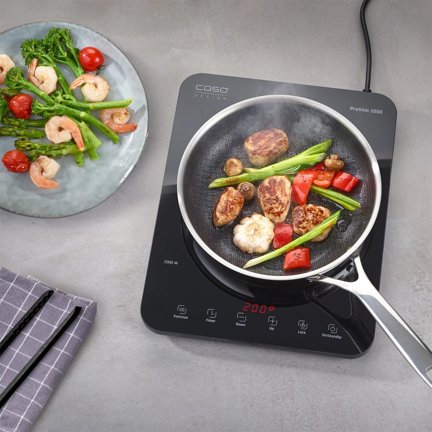 Poêle avec nourriture sur plaque à induction, à côté d'une assiette avec crevettes et légumes.