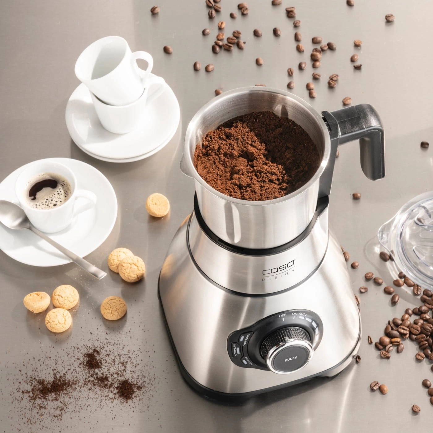 Coffee grinder with coffee, cups, cookies, and coffee beans on a metal surface.