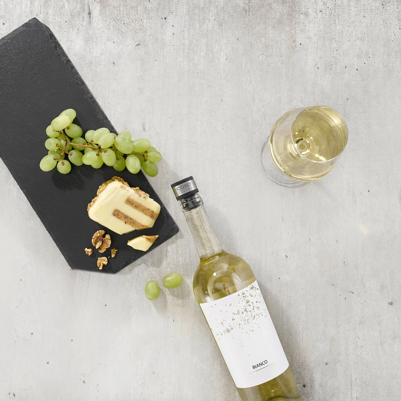 Ein Glas und eine Flasche Weißwein mit Käse, Trauben und Nüssen auf einem Steinbrett.