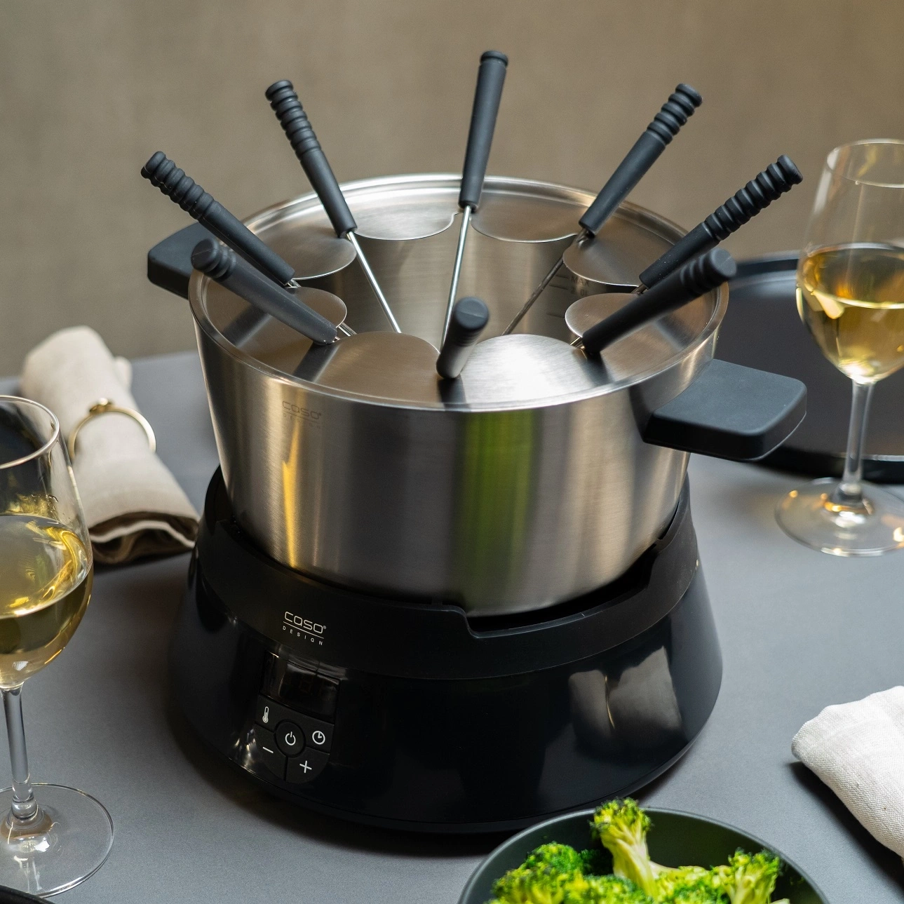 Ein elektrisches Fondue-Set mit acht Gabeln und einem Topf auf einem schwarzen Sockel.
