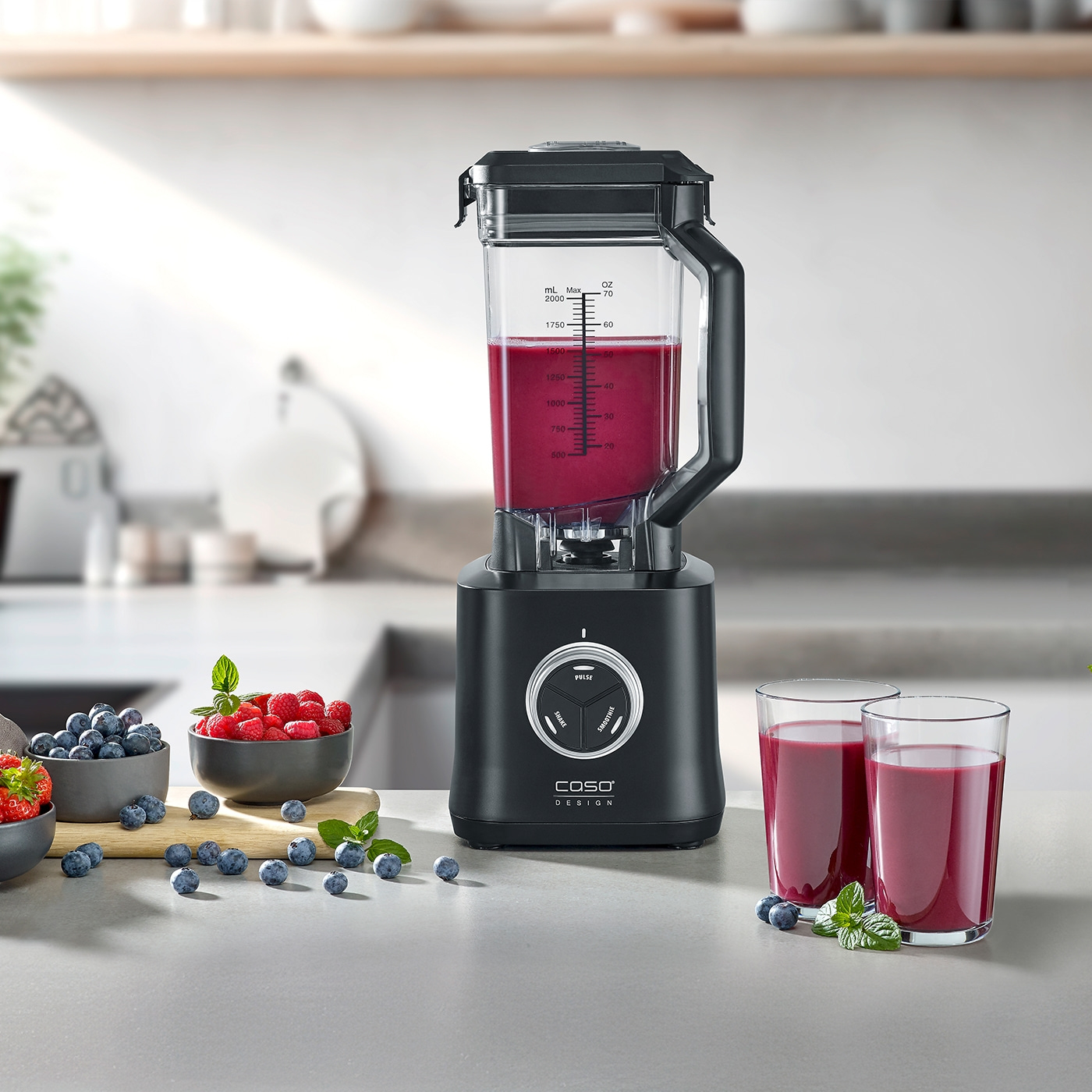 Schwarzer Mixer mit Smoothie, Beeren und Gläsern auf einer Küchenarbeitsplatte.