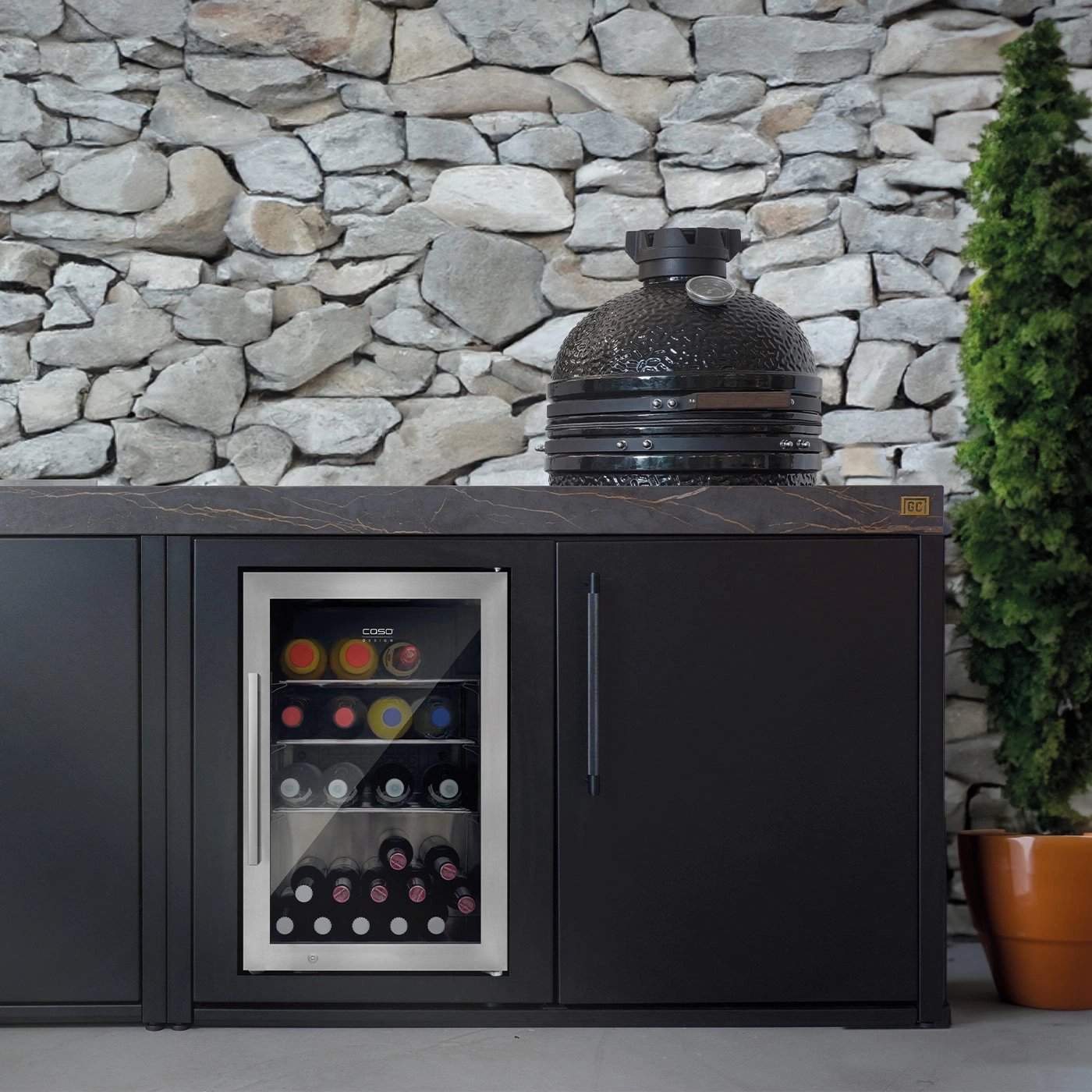 Outdoor-Küche mit Weinkühler, Grill und Steinmauer im Hintergrund.