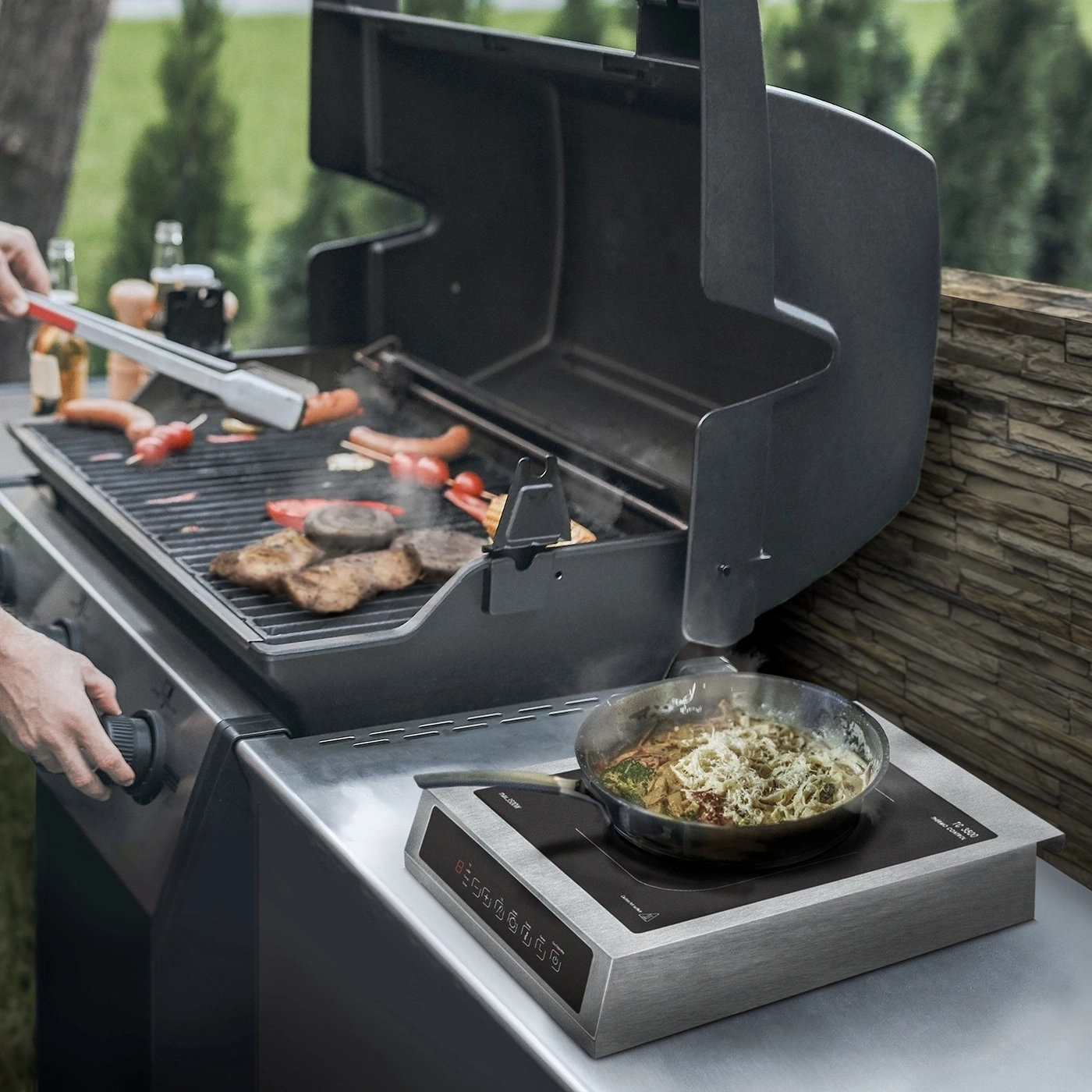 A man grills meat and sausages on a gas grill, with an induction cooktop and a pan next to it.
