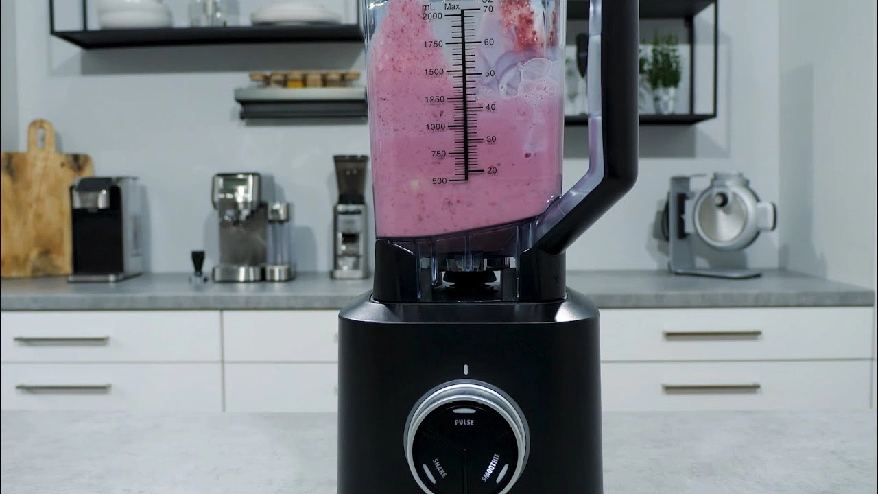 Black blender with pink smoothie in a kitchen with appliances and cabinets.