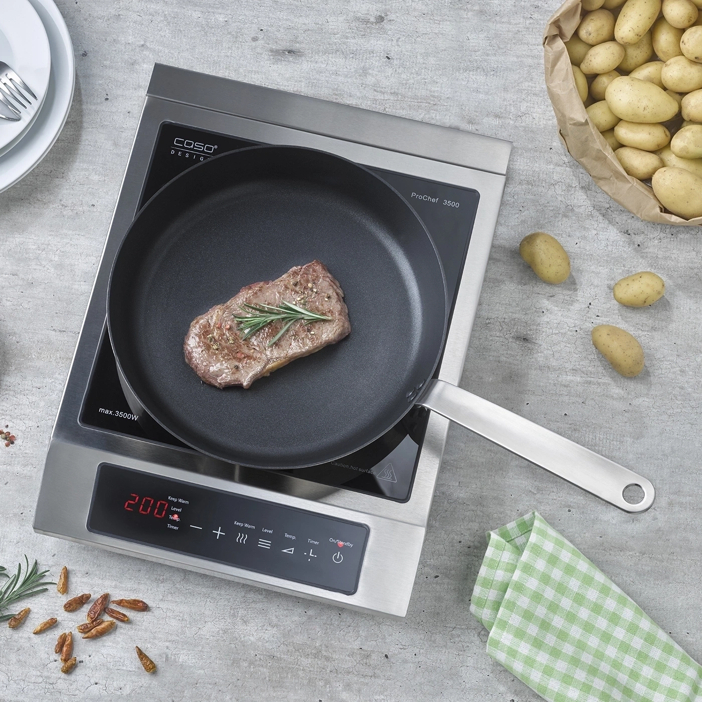 Un steak est frit dans une poêle sur une plaque à induction, avec des pommes de terre et des épices à proximité.
