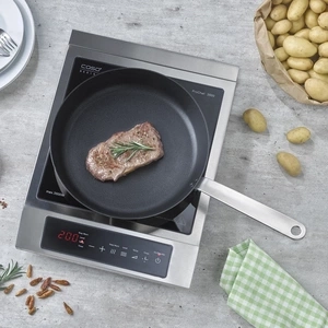 A steak is being fried in a pan on an induction hob, with potatoes and spices nearby.