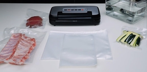 Vacuum sealer with meat, vegetables, and accessories on a white table.