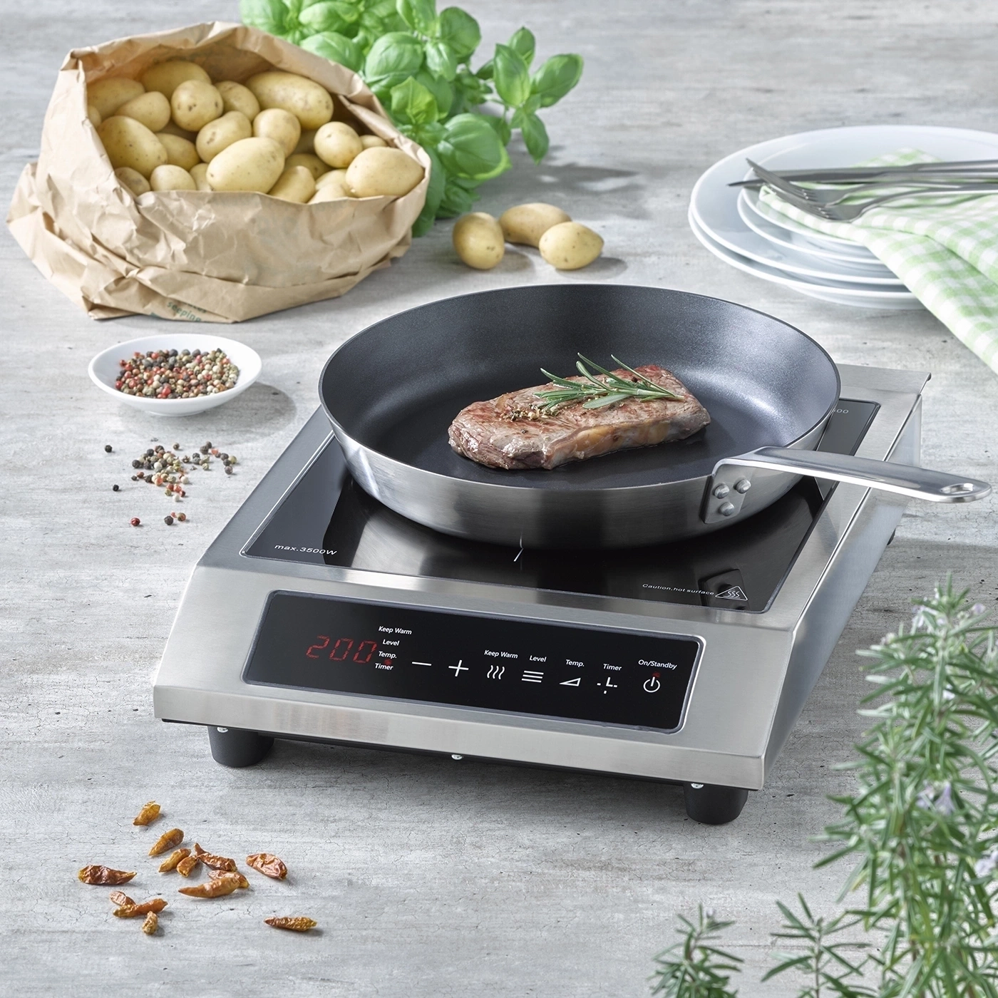 A steak fries in a pan on an induction cooktop, with potatoes and spices nearby.