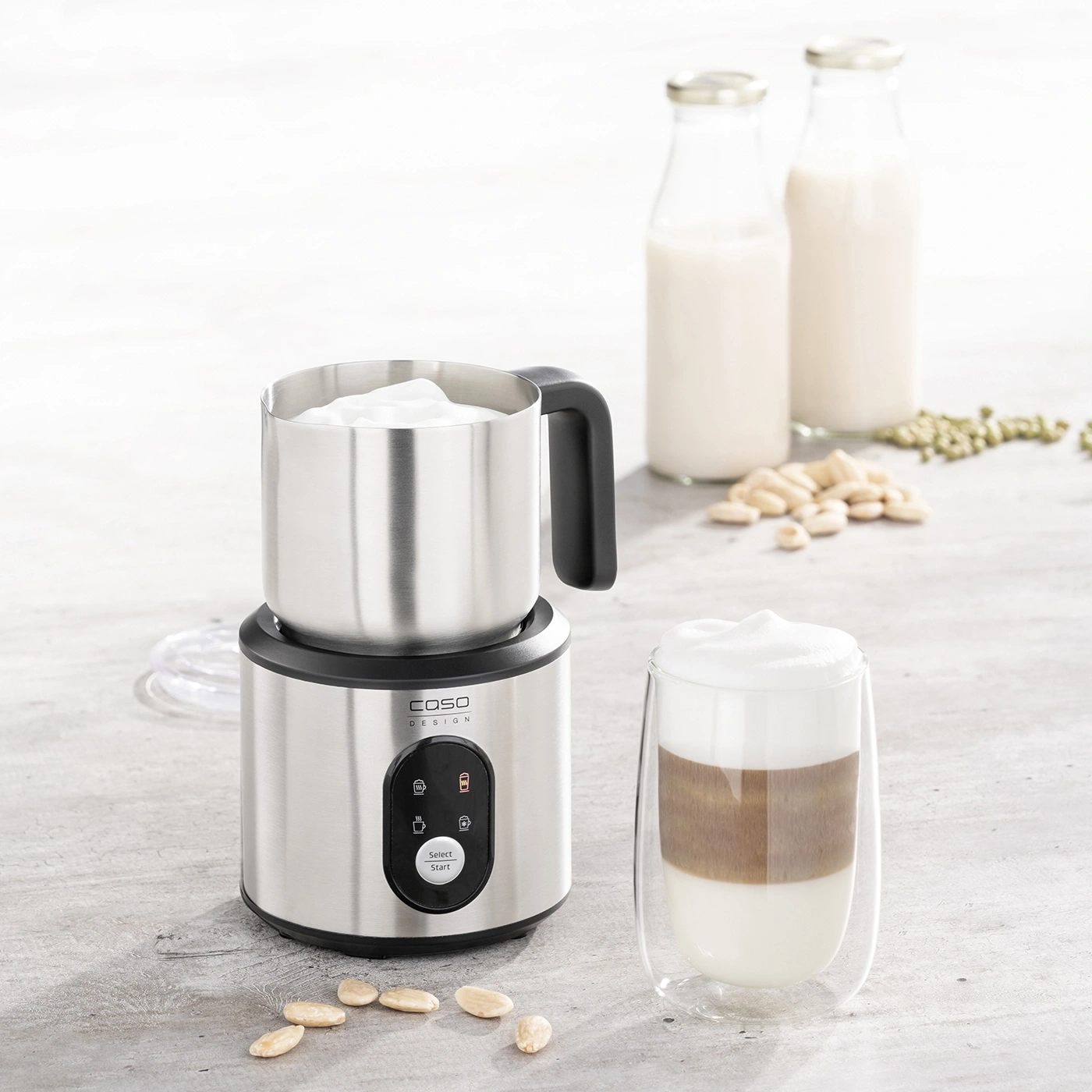 Milk frother with latte macchiato, milk bottles, and almonds on a table.