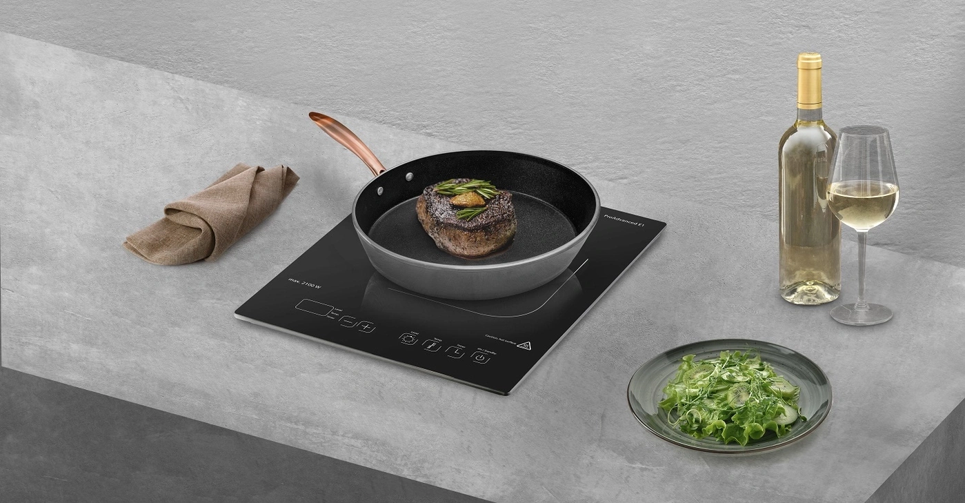 Ein Steak wird in einer Pfanne auf einem Induktionskochfeld gebraten, daneben ein Salat, Wein und ein Glas.